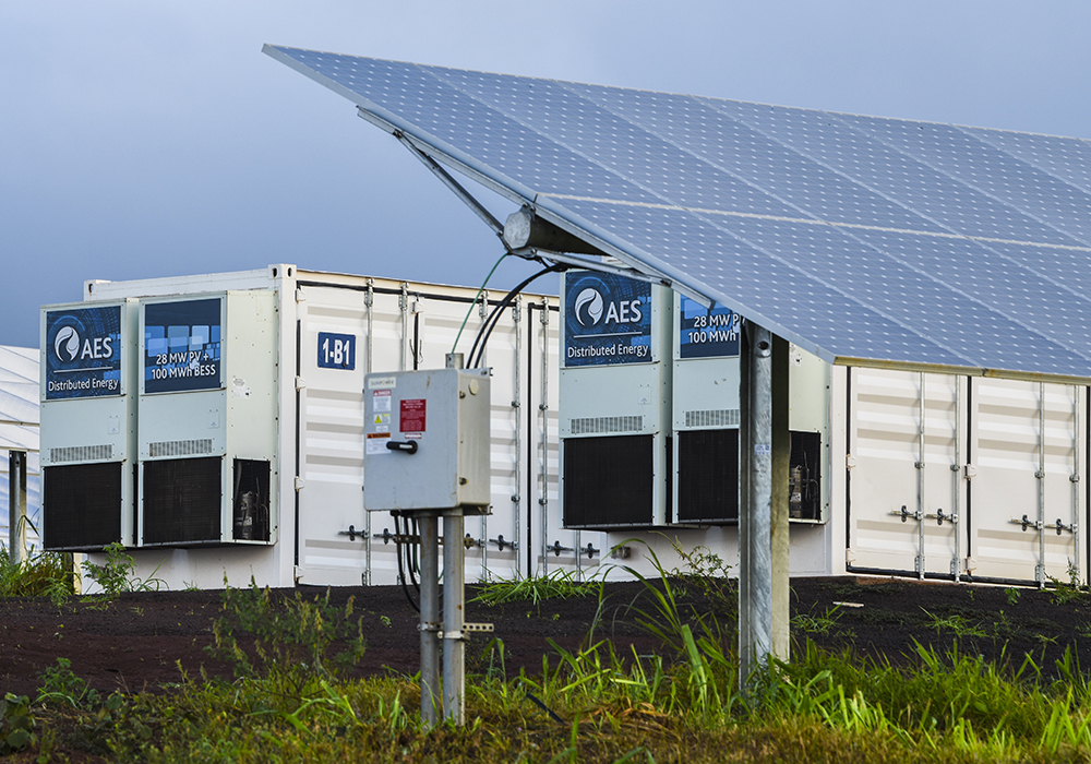 Two rows of intermodal-sized battery enclosures between rows of ground-mounted solar arrays.